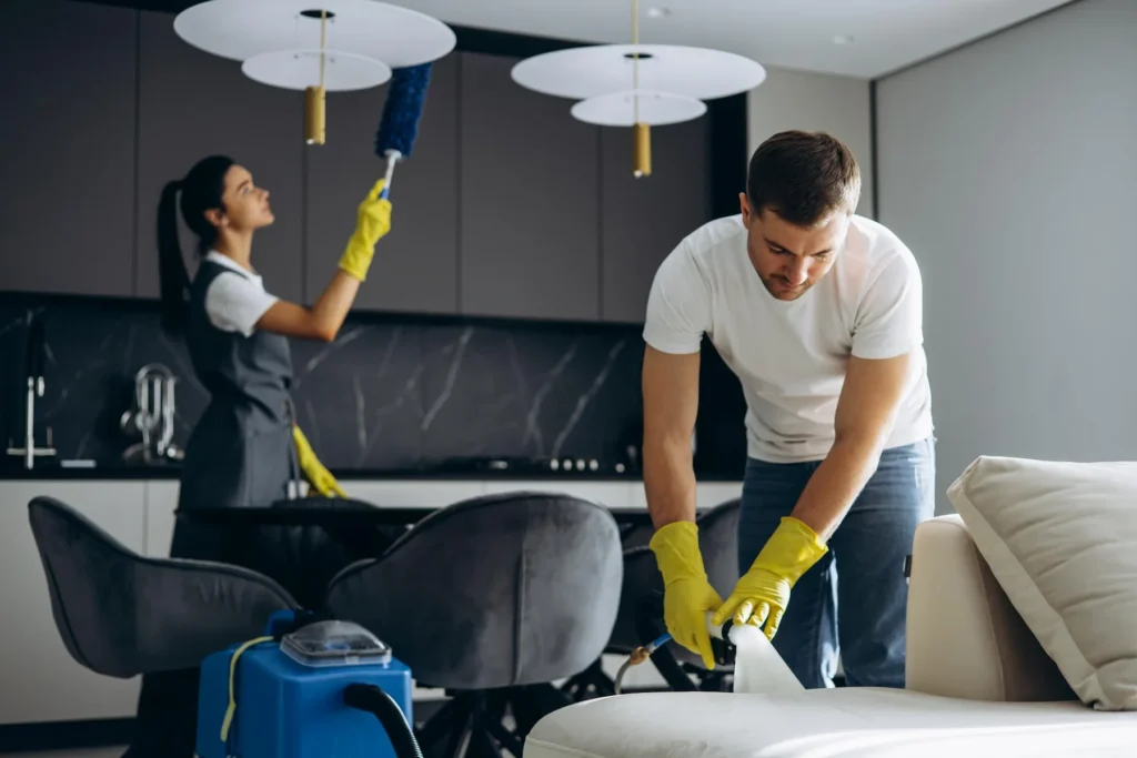 Two professional cleaners in a modern living space, one dusting a high surface and the other cleaning a lower area.