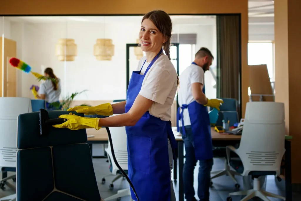 Smiling female cleaner in a blue apron and yellow gloves cleaning an office chair with equipment, with another cleaner in the background.